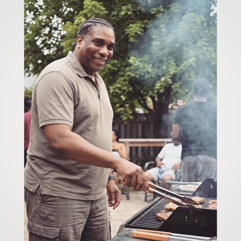 David Elisma souriant au barbecue avec des pinces à la main, devant le gril et des proches réunis dans la cour