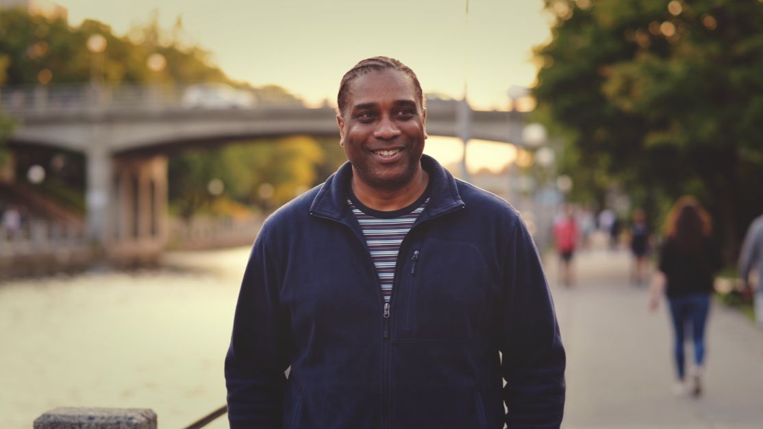 David Elisma souriant sur le sentier du canal Rideau, avec l'eau, un pont et des marcheurs derrière lui