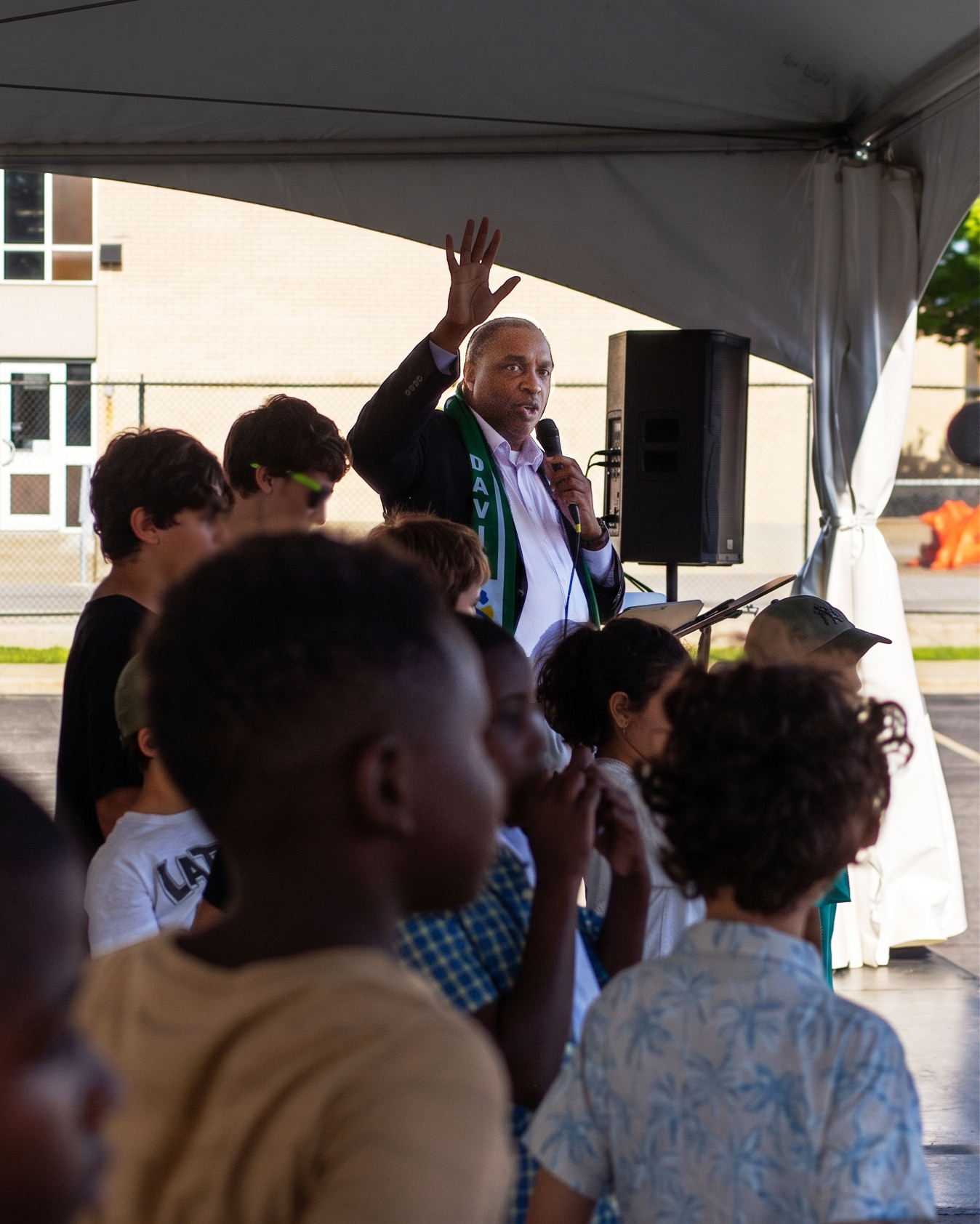 David Elisma speaking into a microphone under a tent in Vanier, one hand raised, with children gathered in front of him at Festi-Mosaïque.