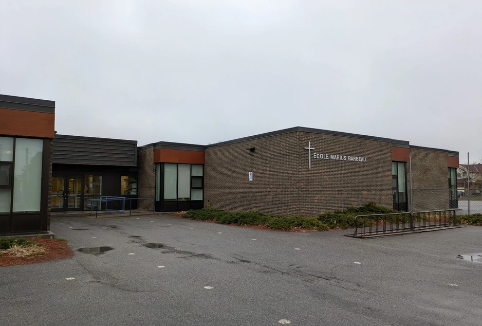 L'École élémentaire catholique Marius-Barbeau vue de sa cour asphaltée : un bâtiment de plain-pied en brique, avec une croix et le nom de l'école sur le mur d'angle.