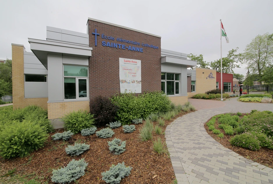 L'entrée de l'École élémentaire catholique Sainte-Anne, dont le nom et la croix se détachent sur un mur de brique, devant des plates-bandes aménagées.
